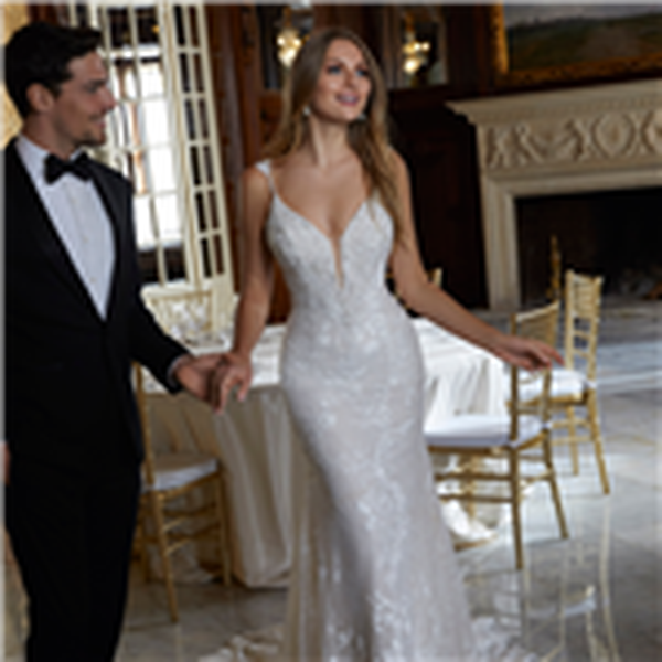 Bride and groom stood in a ballroom near a cream fireplace. Bride wears a strappy lace fit and flare wedding dress with a plunging illusion neckline and scalloped lace hem.
